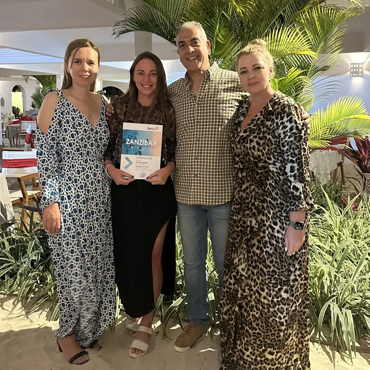 Three smiling girls and one man in front of a palm tree at the Zanzibar Hotel hold a certificate of travel agency Join UP!